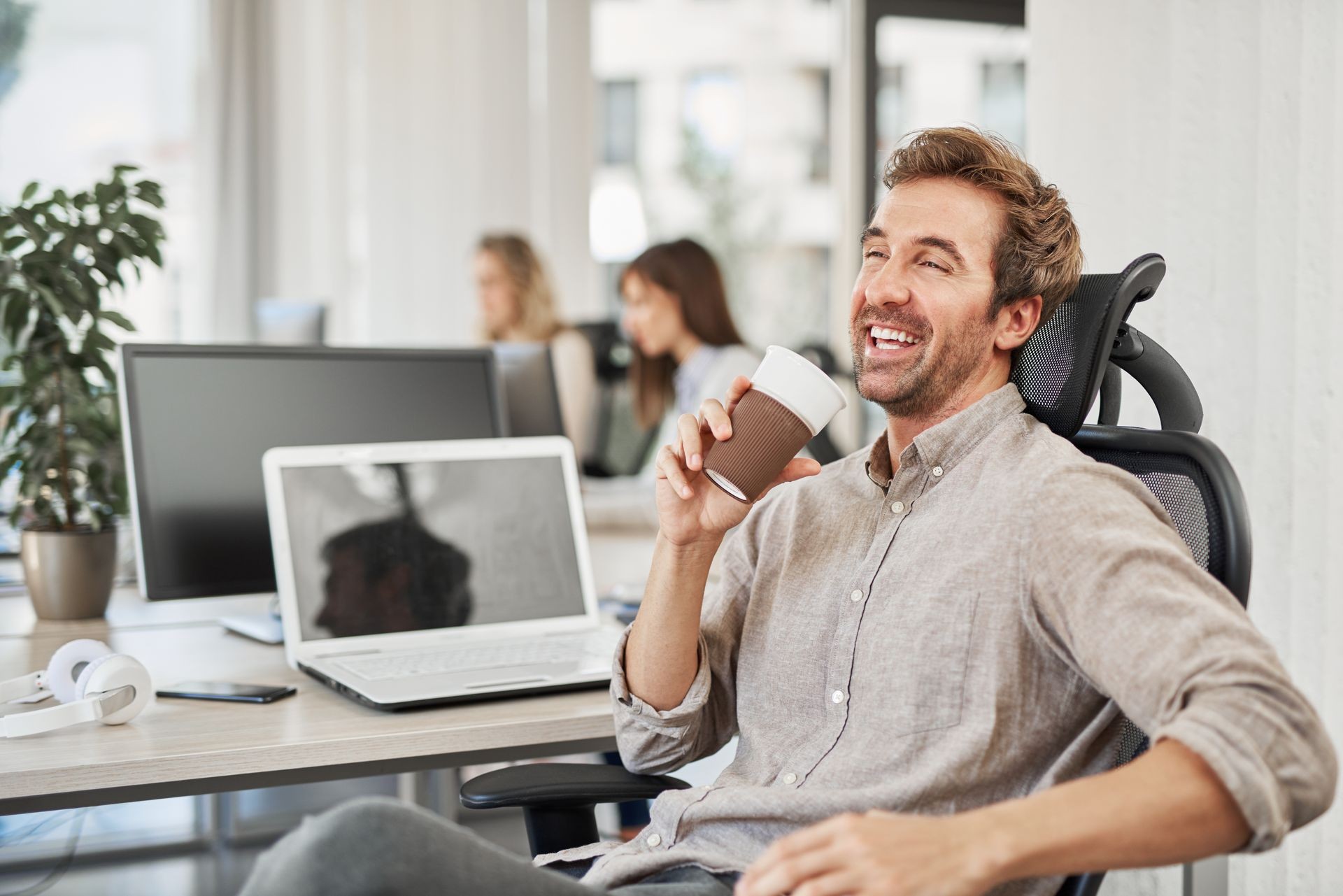 Smiling successful CEO talking to employee and drinking coffee to go while sitting in chair in office. In background workers ar their working stations.